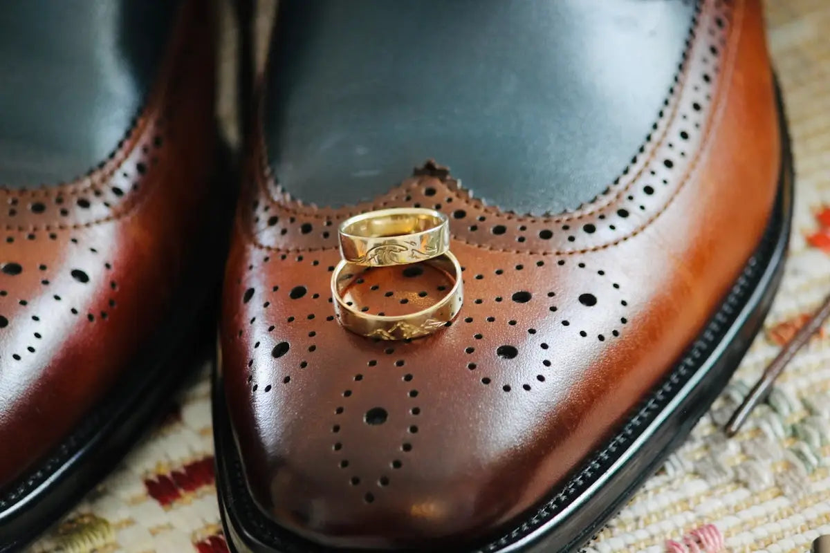 A pair of brown leather brogue dress shoes with black accents and decorative perforations, featuring two gold wedding bands resting on the toe.