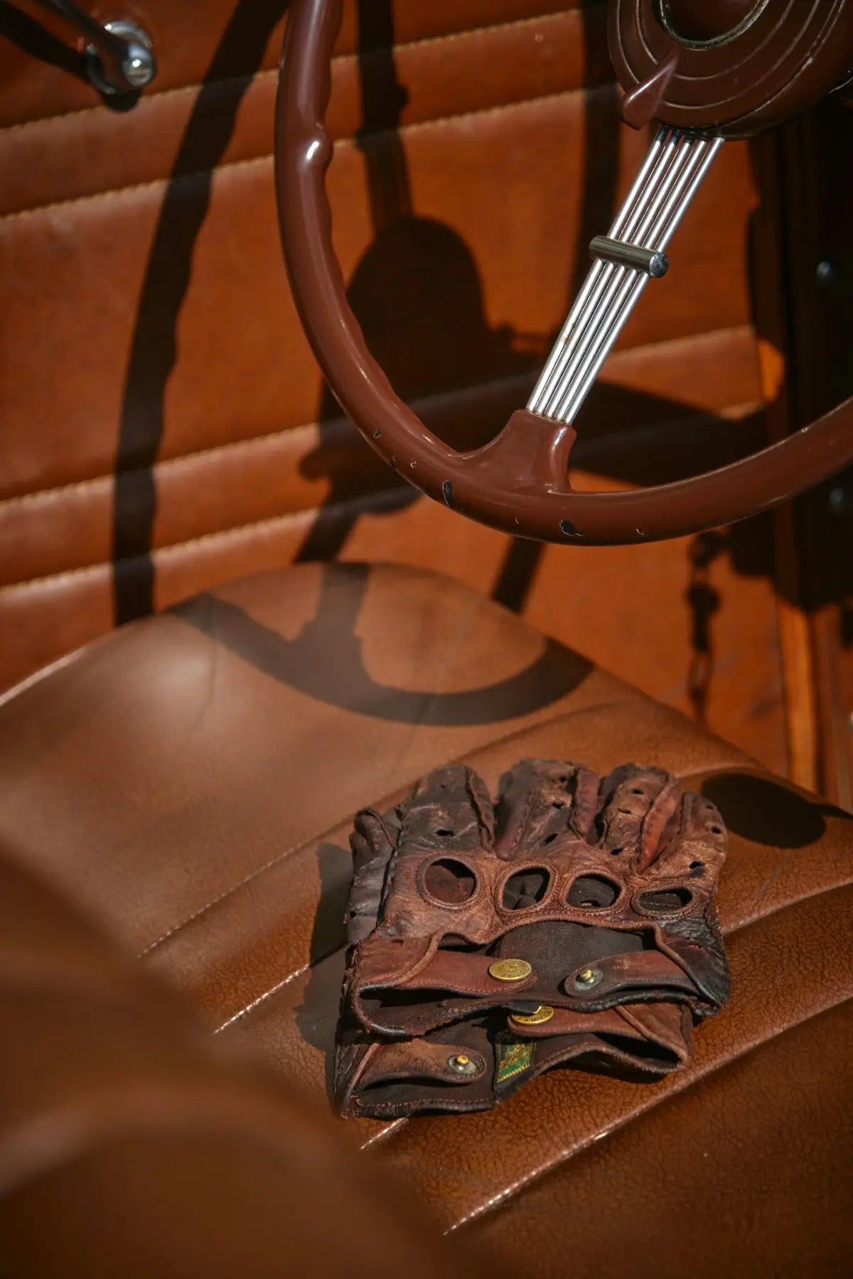 A pair of well-worn brown leather driving gloves with brass rivets and finger holes, resting on a matching leather car seat.